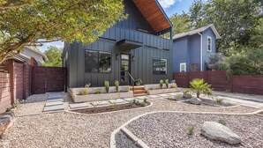 A modern house with a dark exterior, a wooden deck, and a landscaped garden with gravel pathways and small plants.