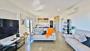 Una sala de estar moderna con un sofá gris, una mesita de centro y un televisor. Se observa una cocina al fondo con armarios de madera y un comedor.