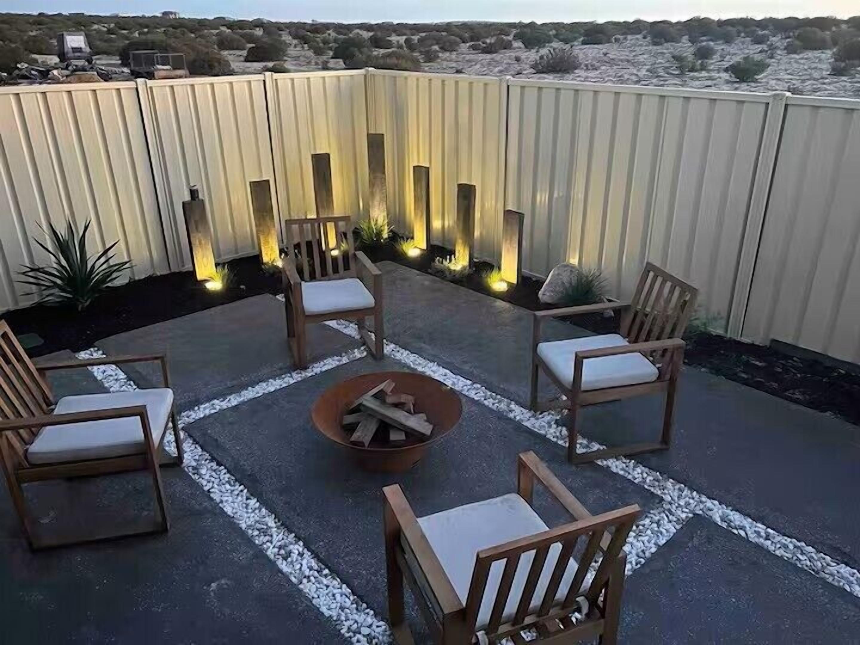 A patio with four wooden chairs and a central fire pit, surrounded by a low wall with vertical light fixtures.