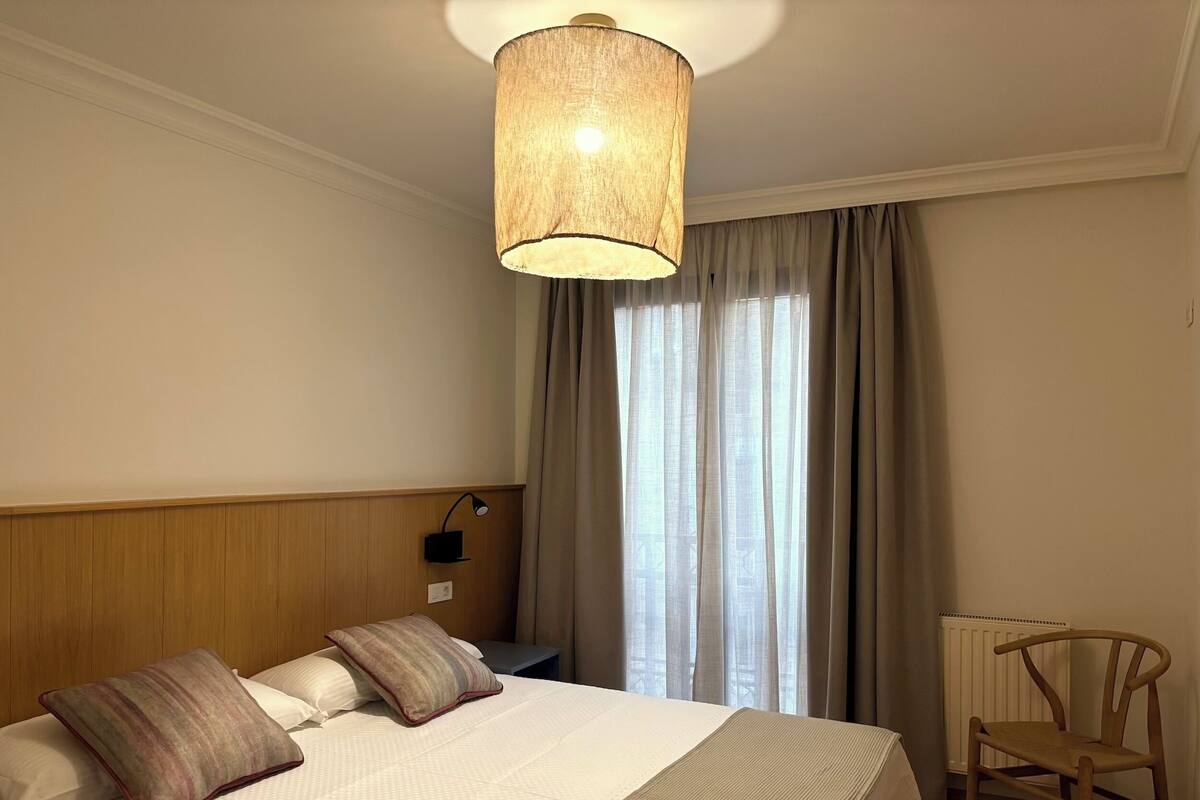 A hotel room with a bed, a desk, a chair, a lamp, and a window with curtains.