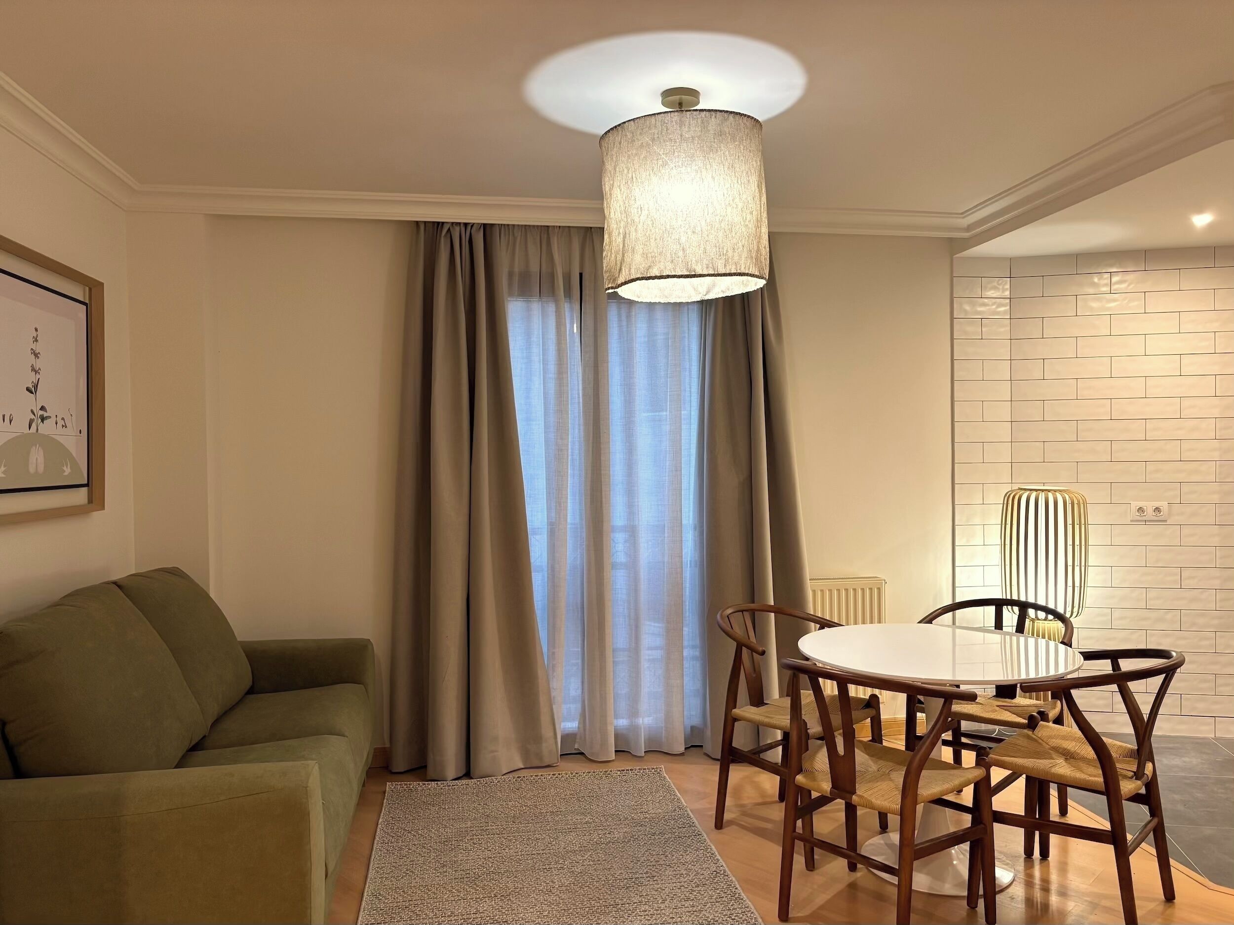 Una sala de estar con un sofá verde, una mesa de comedor redonda con sillas de madera, una lámpara colgante y una ventana con cortinas.