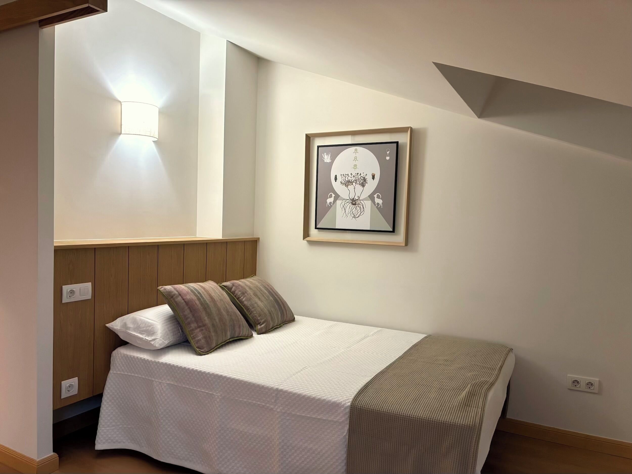 A small, neatly arranged bedroom with a bed, a wall-mounted light, and a framed artwork.