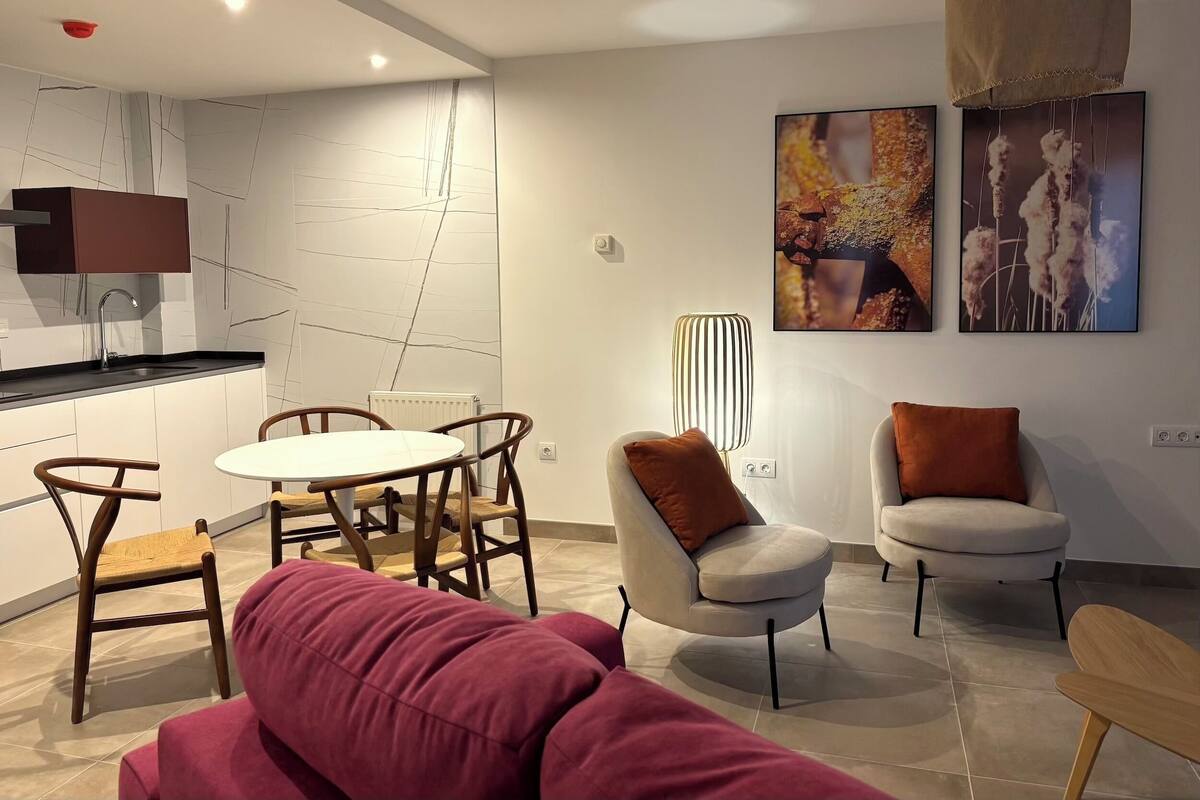 A modern living room with a sofa, two armchairs, a dining table with chairs, a kitchen area, and artwork on the walls.