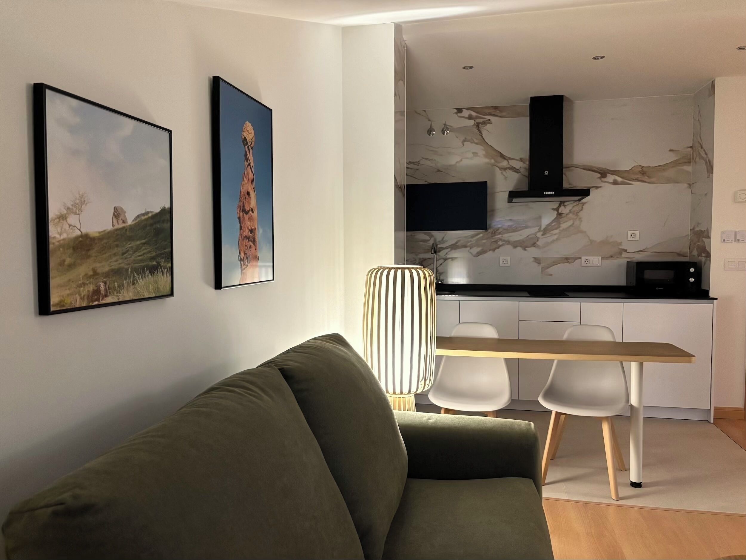 Una sala moderna con un sofá verde, una mesa de comedor con dos sillas y una cocina con salpicadero de mármol y una campana extractora negra.