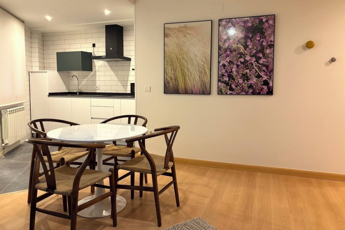 A modern kitchen with white tiles, a black range hood, and a white countertop. There is a dining area with a round table and four chairs. Two framed pictures are on the wall.