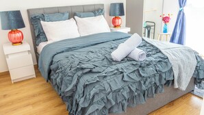 A neatly made bed with a grey ruffled bedspread, two bedside lamps, and a small table with a vase of flowers. - Cozy, Spacious condo with balcony and beautiful serene mountain views. (Cluses)
