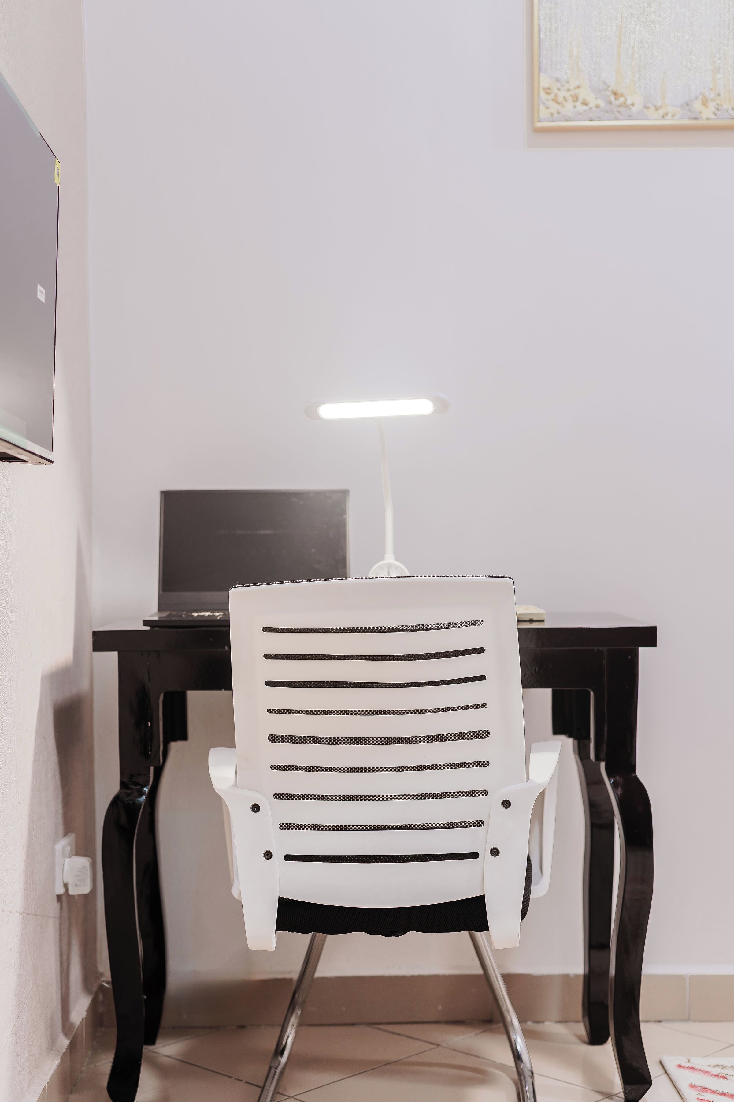 A white office chair with a backrest and armrests, positioned in front of a desk with a computer monitor and a white desk lamp.