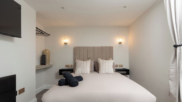 Une chambre à coucher avec un grand lit, deux tables de chevet, un téléviseur fixé au mur et une étagère ornée d’objets décoratifs.
