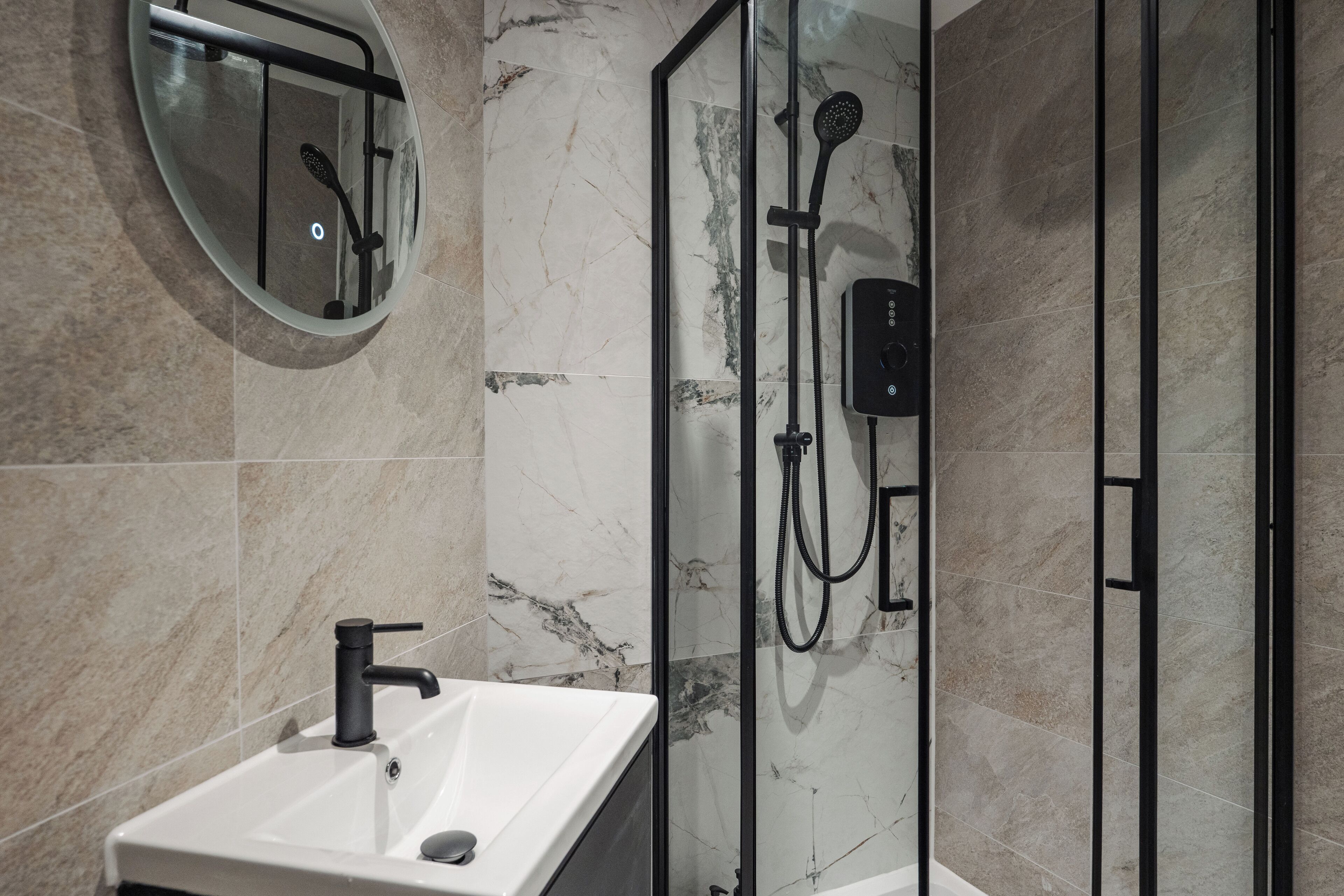 A modern bathroom with a glass-enclosed shower, a round mirror, and a white sink.