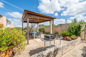 Une terrasse extérieure couverte, équipée d’un barbecue, d’une table, de chaises et de plantes en pot.