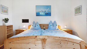 Uma cama de madeira com cabeceira, uma mesa de cabeceira com abajur, uma prateleira na parede com uma planta e um quadro de um veleiro.
