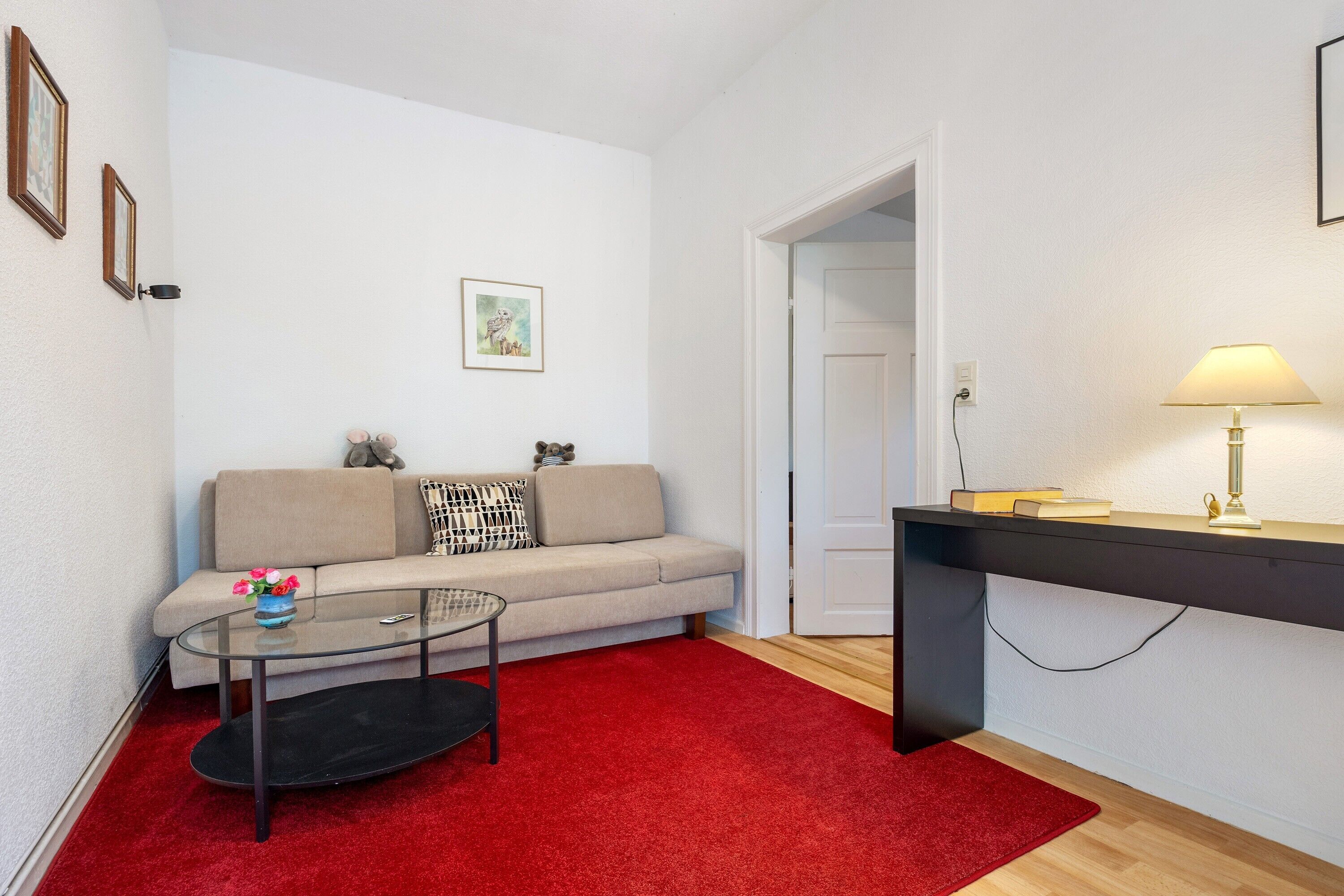 Een woonkamer met een bank, een salontafel, een rood tapijt, een lamp en een deur die naar een andere kamer leidt.