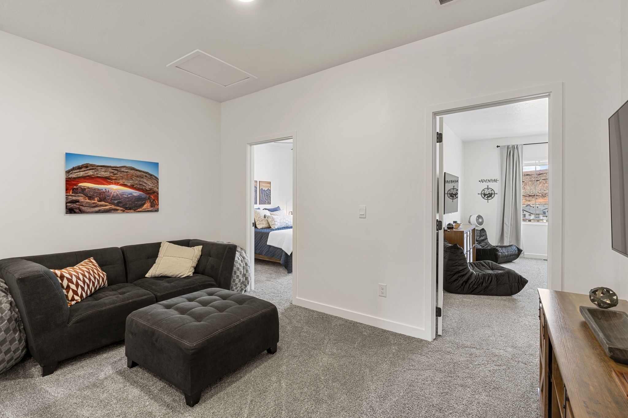 A modern living room with a dark grey sofa, a matching ottoman, and a large painting of a canyon. There is a doorway leading to a bedroom with a bed and a bathroom visible.