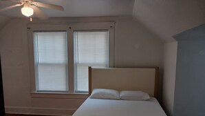Une chambre à coucher avec un lit, deux fenêtres équipées de stores, un ventilateur de plafond et un plancher en bois.