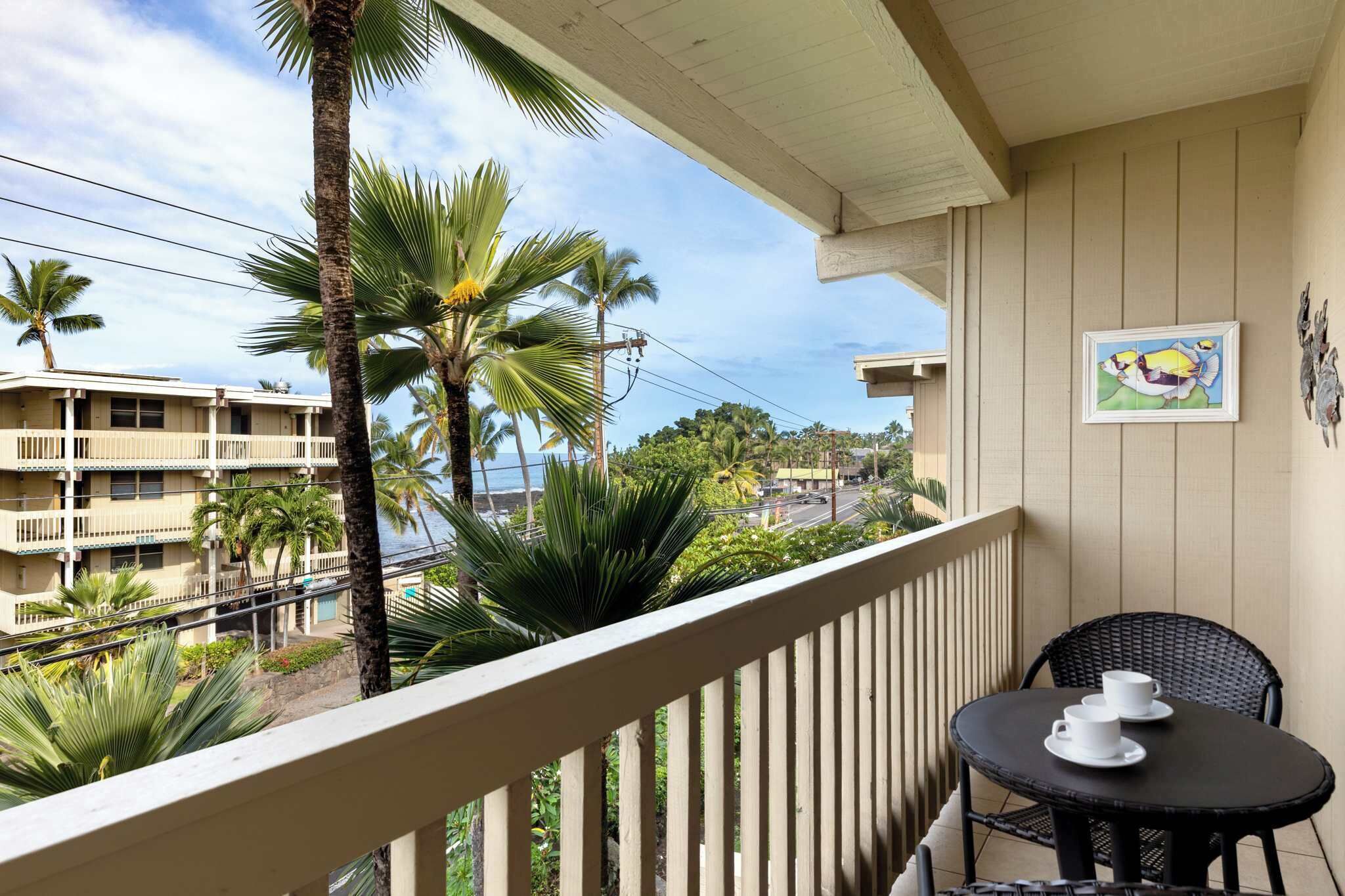 A balcony with a view of palm trees and buildings, a small table with cups, and a framed picture on the wall.