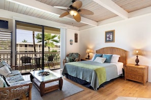 A room with a bed, a table, chairs, a ceiling fan, and a balcony with a view of palm trees.