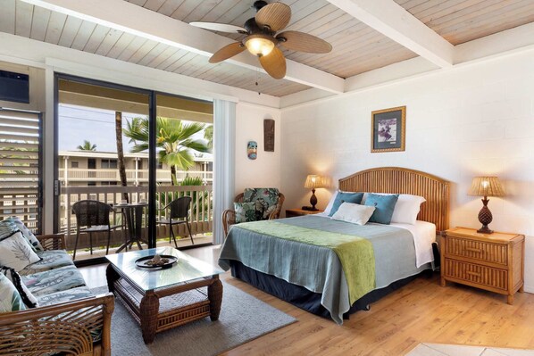 Ein Zimmer mit einem Bett, einem Tisch, Stühlen, einem Deckenventilator und einem Balkon mit Blick auf Palmen.
