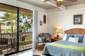 A hotel room with a bed, a chair, a table, a lamp, and a view of palm trees.