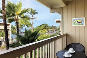 A balcony with a view of the ocean, palm trees, and a painting on the wall.