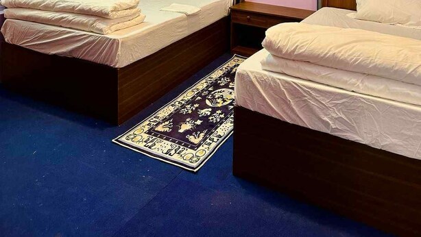 Hotellihuone, jossa on kaksi sänkyä, puinen pääty, yöpöytä, sängyn vieressä oleva pöytä, kuvioitu matto ja kattovalaisin.