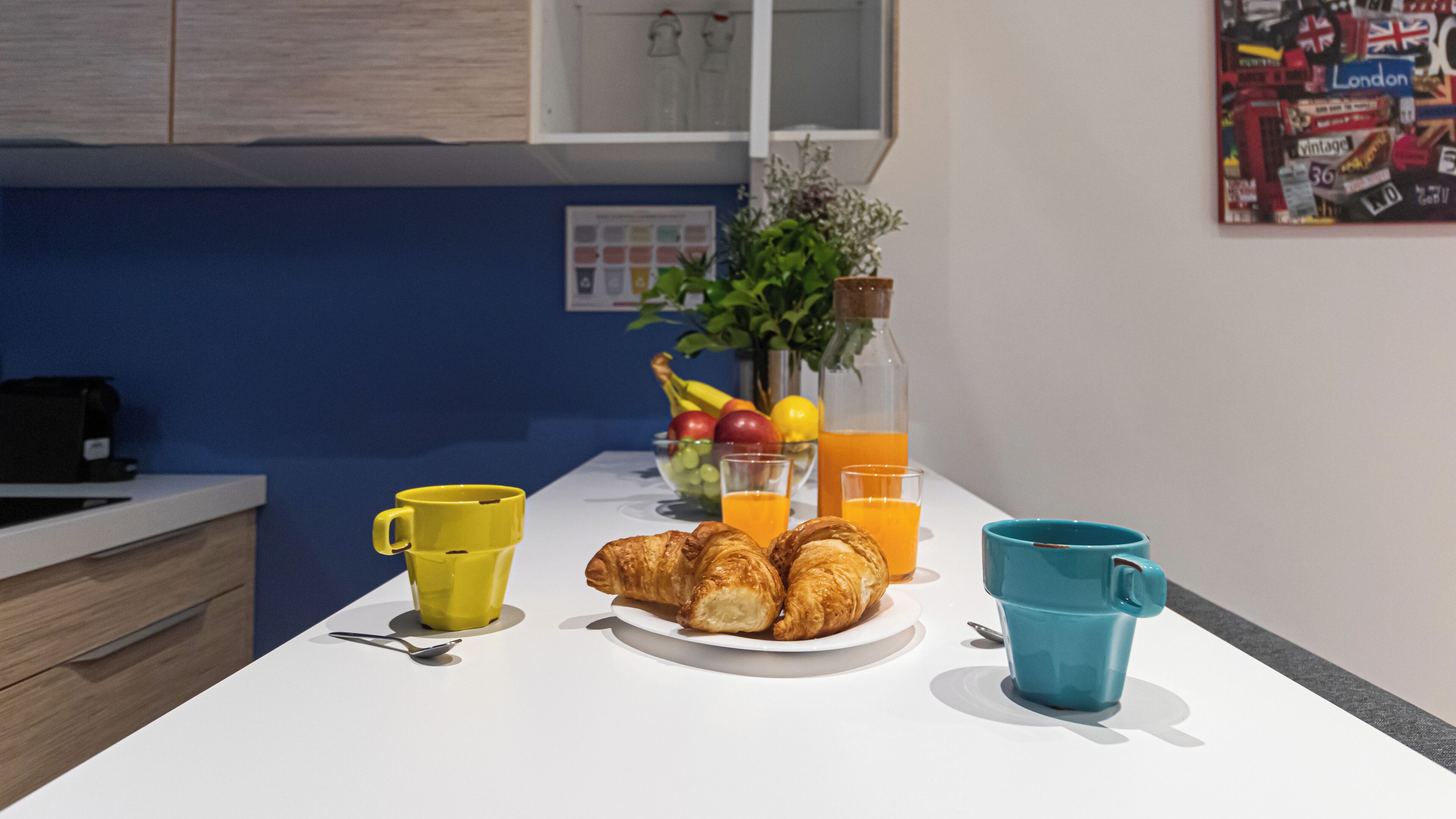 Een keukentoog met een bord croissants, twee bekers, een fruitschaal en een fles sinaasappelsap.