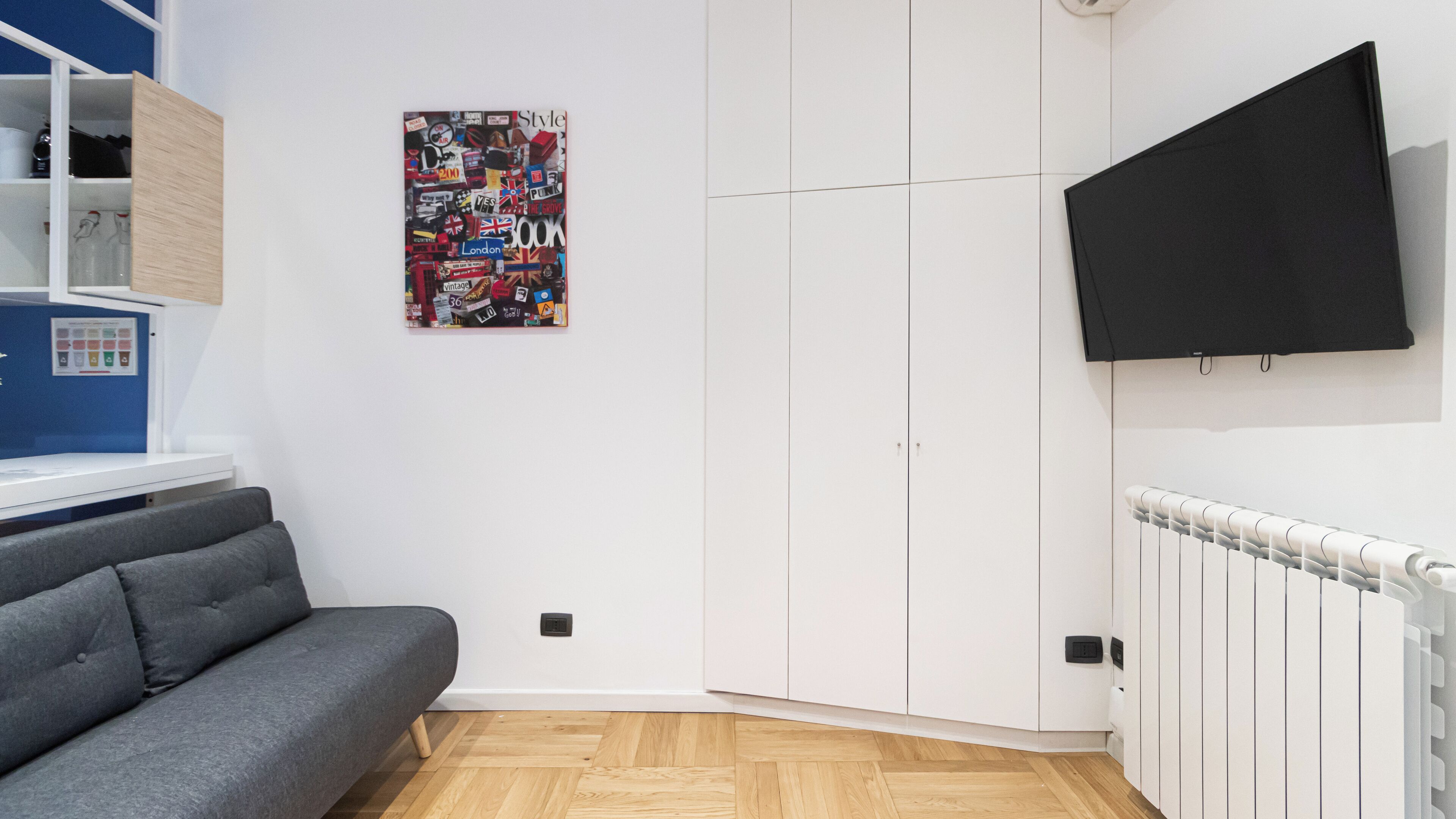 Un salon moderne avec un canapé gris, une armoire blanche, un téléviseur fixé au mur et une affiche colorée accrochée au mur.