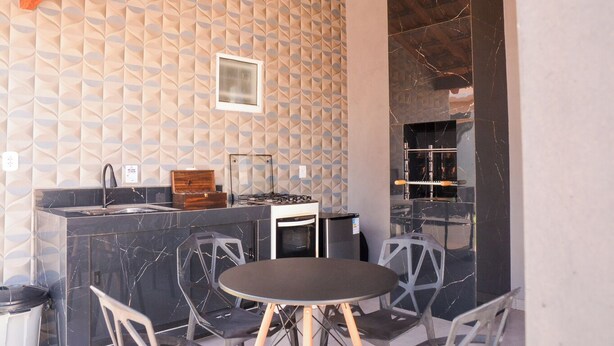 An outdoor kitchen area with a marble countertop, a sink, and a stove. There are two chairs and a table in front. The wall has a geometric pattern. - Casa com Garagem e Churrasqueira em Tima Regi o (Olímpia)