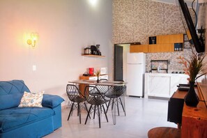 A compact living space with a blue sofa, a dining table with chairs, a kitchen area with a refrigerator, and a wall-mounted light fixture. - Casa com Garagem e Churrasqueira em Tima Regi o (Olímpia)