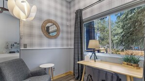 Une pièce avec un mur orné d’un motif à carreaux, un miroir rond, un bureau avec une lampe, une chaise et une vue sur les arbres à l’extérieur.