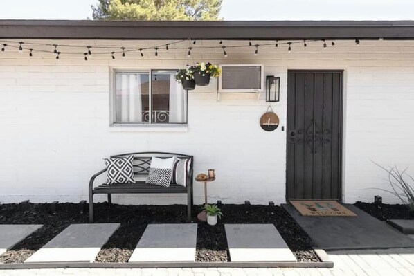 A white house with a black bench, a door, and a window with a hanging plant.