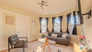 Una sala de estar con un sofá, una mesa de centro y una silla. Se ve una puerta y una ventana con cortinas.