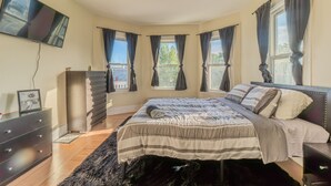 Un dormitorio con cama, cómoda, televisor y ventanas con cortinas.