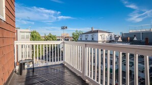 Una terraza de madera con barandilla blanca, con vistas a una zona residencial con casas y una calle.