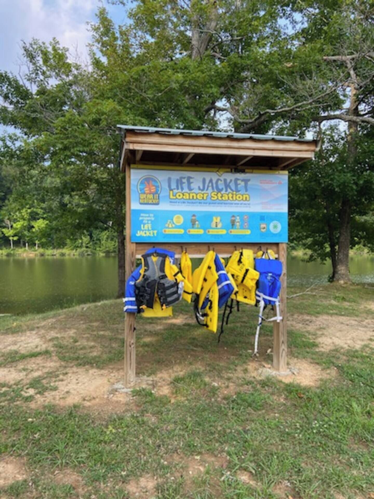 Un poste de distribution de gilets de sauvetage, avec un panneau indicateur et des gilets de sauvetage suspendus à une structure en bois, situé près d’un lac.