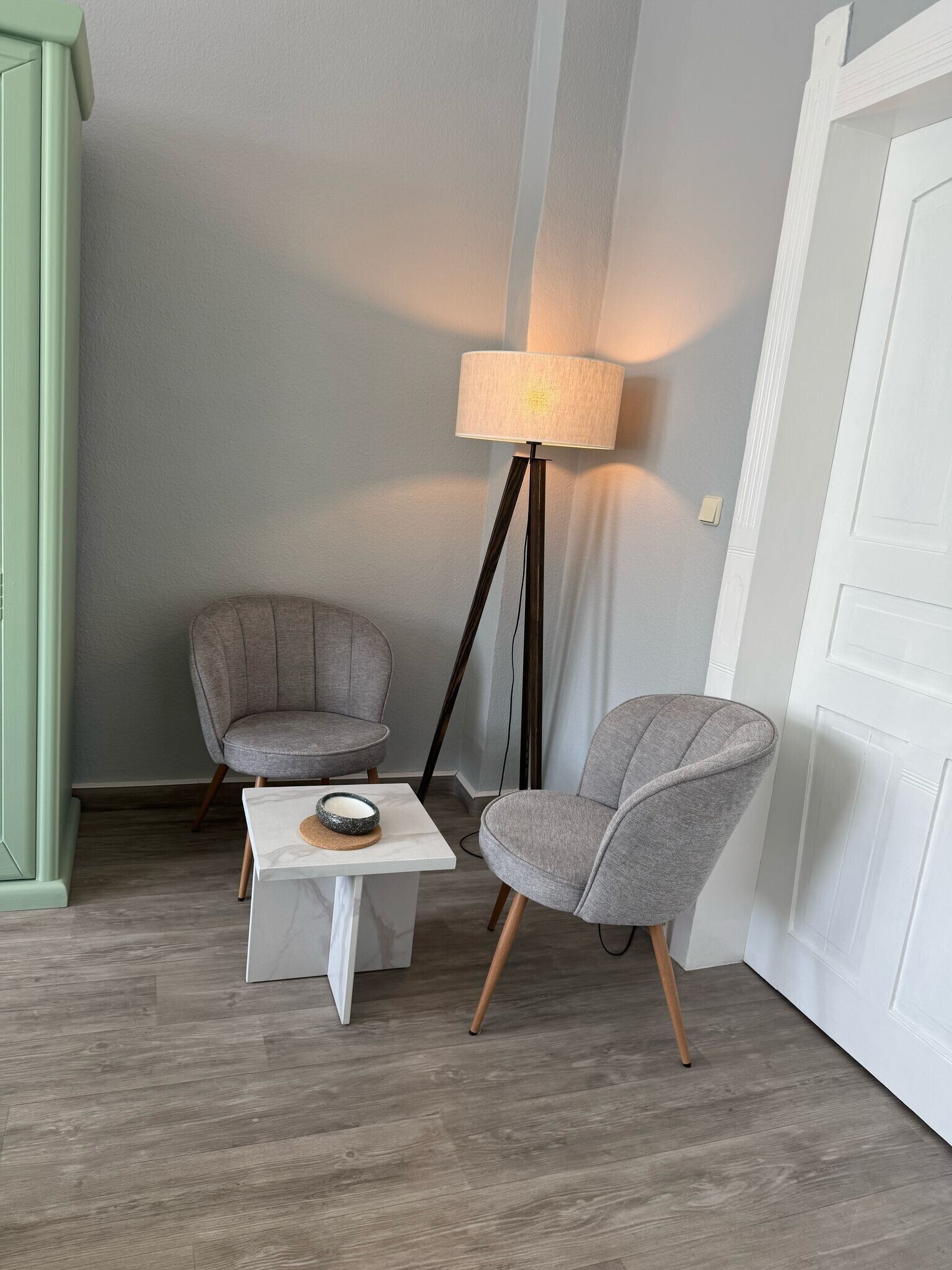 Une pièce avec deux fauteuils gris, une petite table blanche sur laquelle se trouve un bol, un lampadaire et une porte blanche.