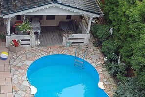 Une piscine avec une terrasse en bois, un espace salon couvert et un chemin en pierre.