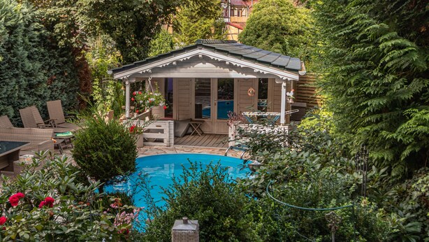 Een overdekt buitenzwembad met een houten terras, omringd door weelderige begroeiing en een klein paviljoen.