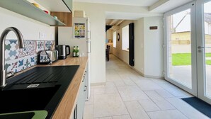 A modern kitchen with a black countertop, stainless steel sink, and a refrigerator with a digital display. A hallway leads to a glass door entrance. - Les Chatons - Gite au coeur de la campagne (Fresville)