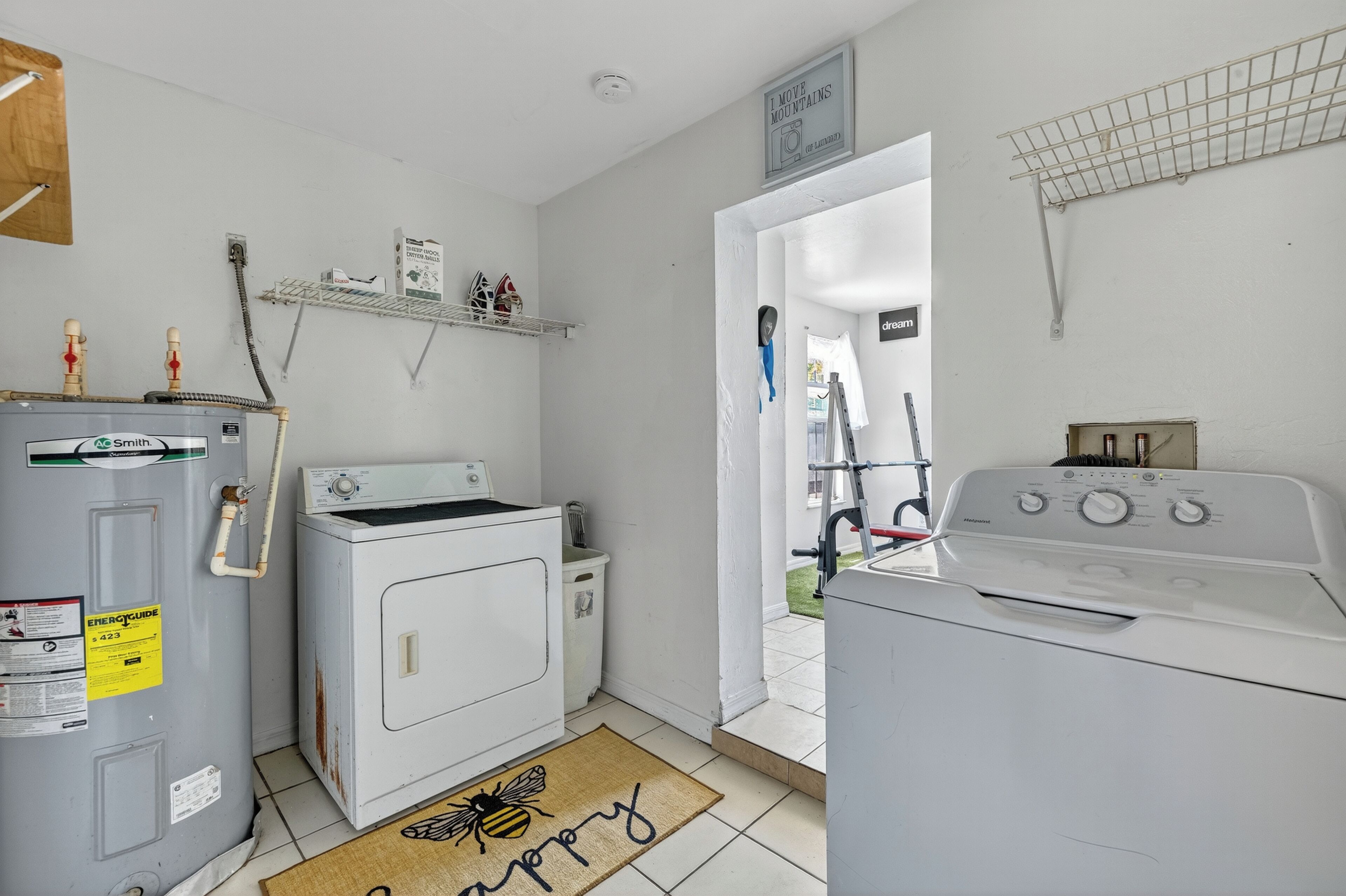 A laundry room with a washer, dryer, and water heater. There is a shelf with cleaning supplies and a sign above the door.