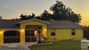 A yellow single-story house with a garage, a front porch, and a small garden. - Spacious home with 7 beds, home gym, office with oversized backyard. (Fort Myers)