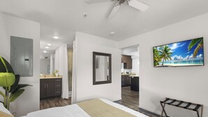 A hotel room with a bed, a ceiling fan, a TV, a mirror, and a view of the kitchen and bathroom.