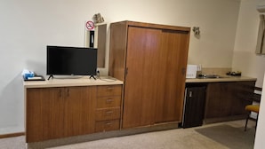 Quarto compacto com guarda-roupa de madeira, televisão de tela plana, pequena cozinha com micro-ondas e pia.