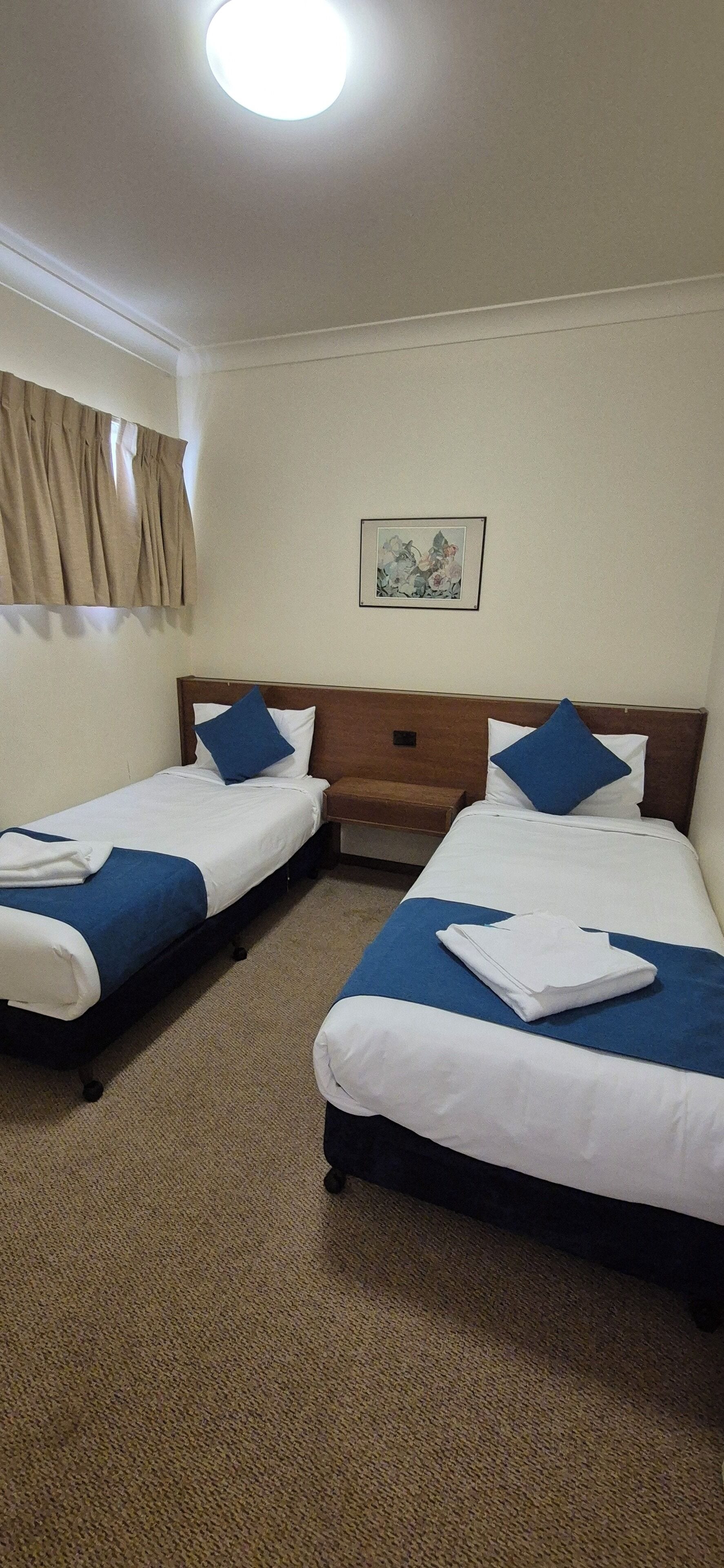 Habitación de hotel con dos camas, cada una con cubrecama azul y blanco, y una cama individual con cubrecama azul y blanco. Hay un cuadro enmarcado en la pared sobre las camas.