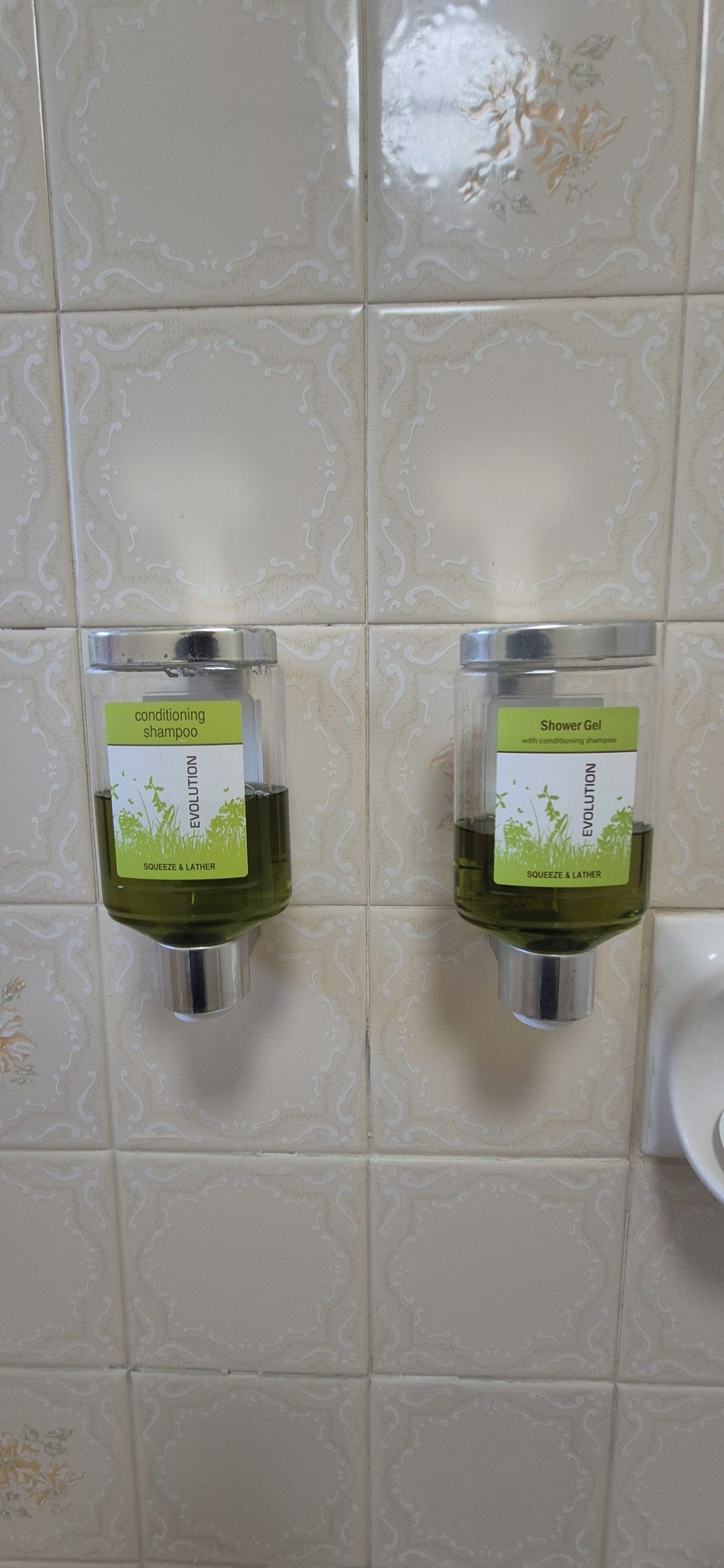 Two dispensers of shower gel and conditioning shampoo mounted on a tiled wall.