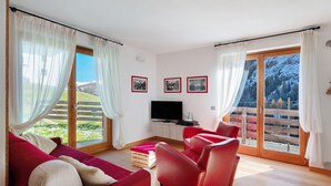 A modern living room with a sofa, red armchairs, a TV, and a balcony with a mountain view. - Apartment 'Sunny Arabba' with Mountain View, Private Garden and Wi-Fi (Arabba)