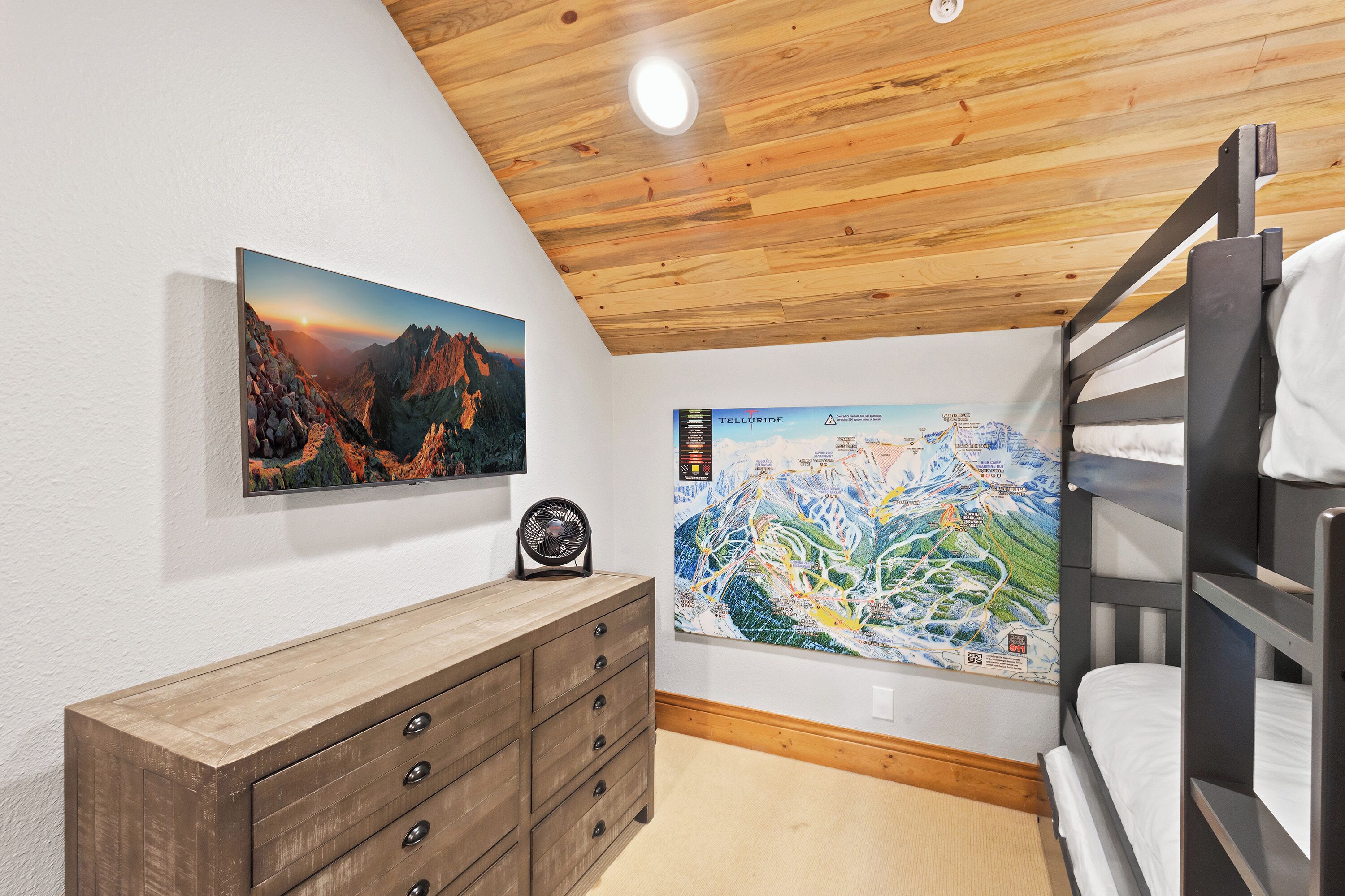 A room with a bunk bed, a wooden dresser, a framed mountain landscape, and a ski resort map.