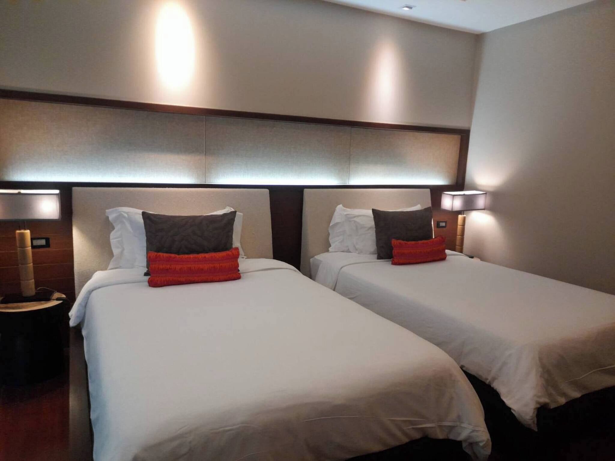 A hotel room with two beds, each with white bedding and a red accent pillow. There are two bedside lamps and a nightstand with a lamp.