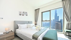 A modern hotel room with a large bed, a bedside table with a lamp, a bench, and a view of a city skyline through a large window. - Burj Khalifa View - 2 BR - PS5 - Pool (Dubai)