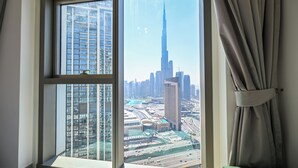 A room with a large window offering a view of a cityscape, including a prominent skyscraper. - Burj Khalifa View - 2 BR - PS5 - Pool (Dubai)