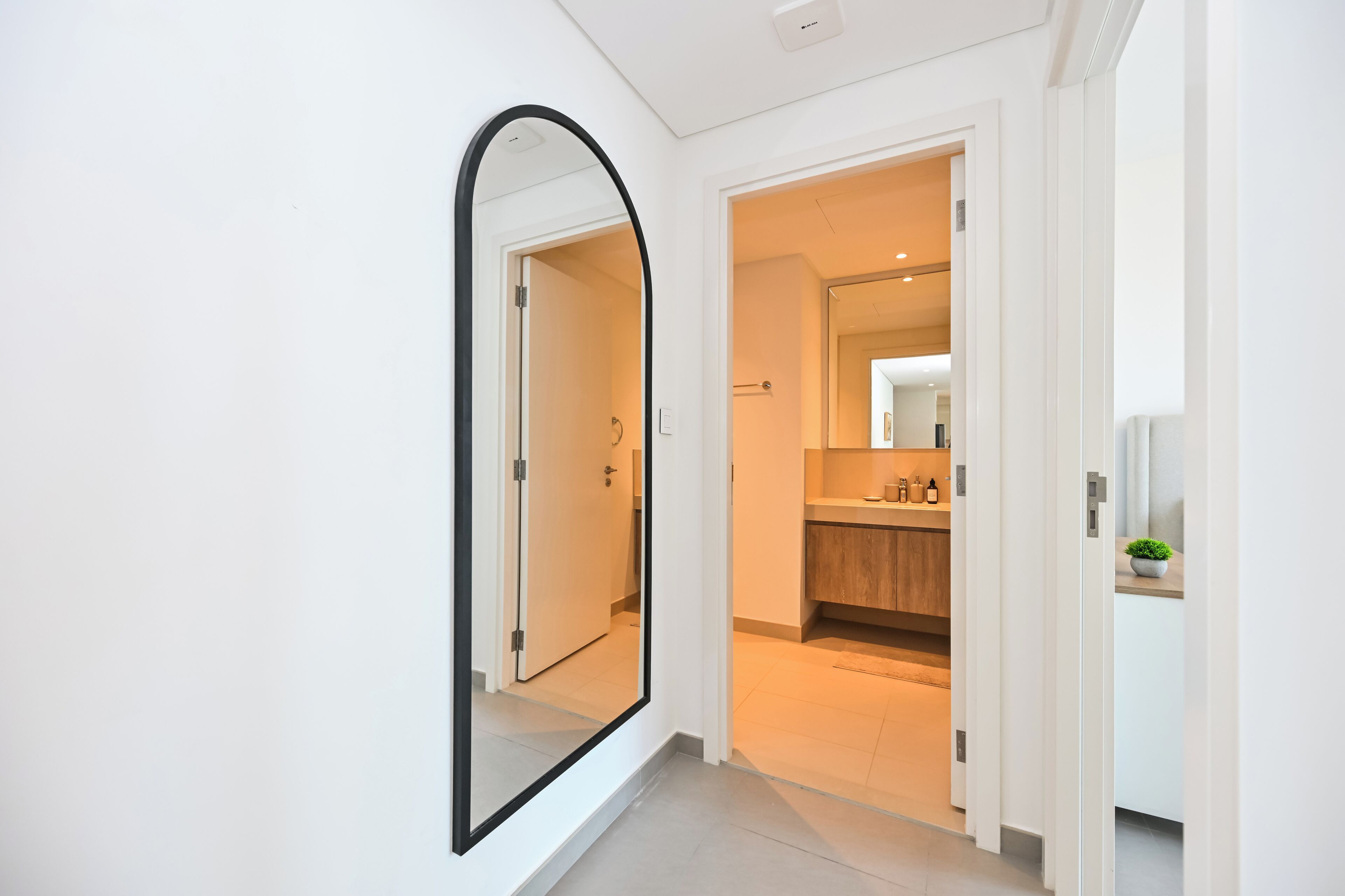 A modern hallway with a large mirror, leading to a bathroom with a wooden vanity and a small plant.